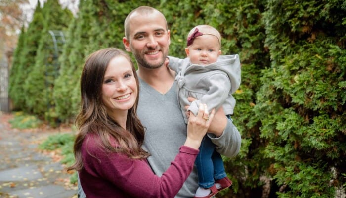 A husband and wife pose with their baby in front of trees.