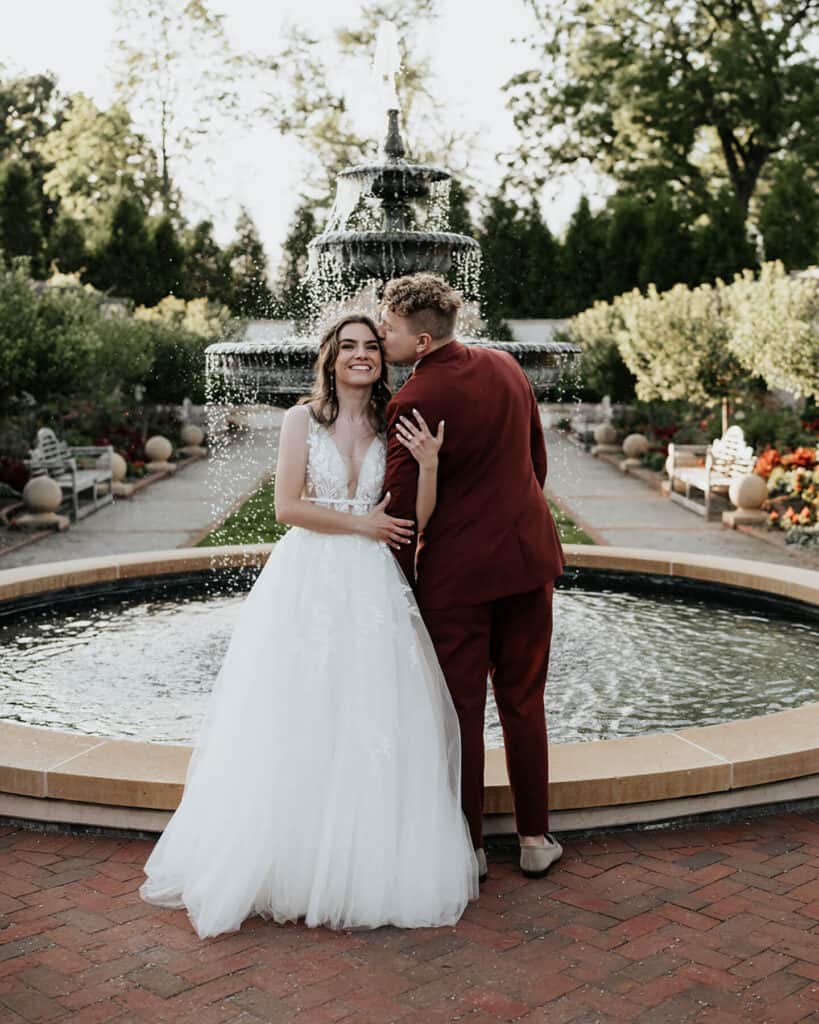 A bride and groom pose in front of the fountain in the Formal Garden.