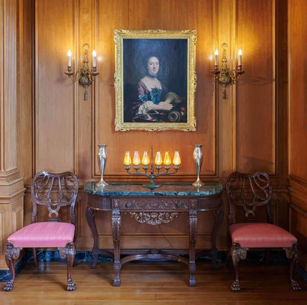 A lamp of six candelabras with iridescent blown-glass shades in an orange hue stand on a credenza surrounded by other décor.