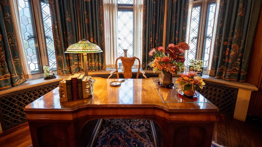 A desk in the Library features mauve, orange, green and teal floral arrangements that match the Tiffany lamp and draperies in the background.