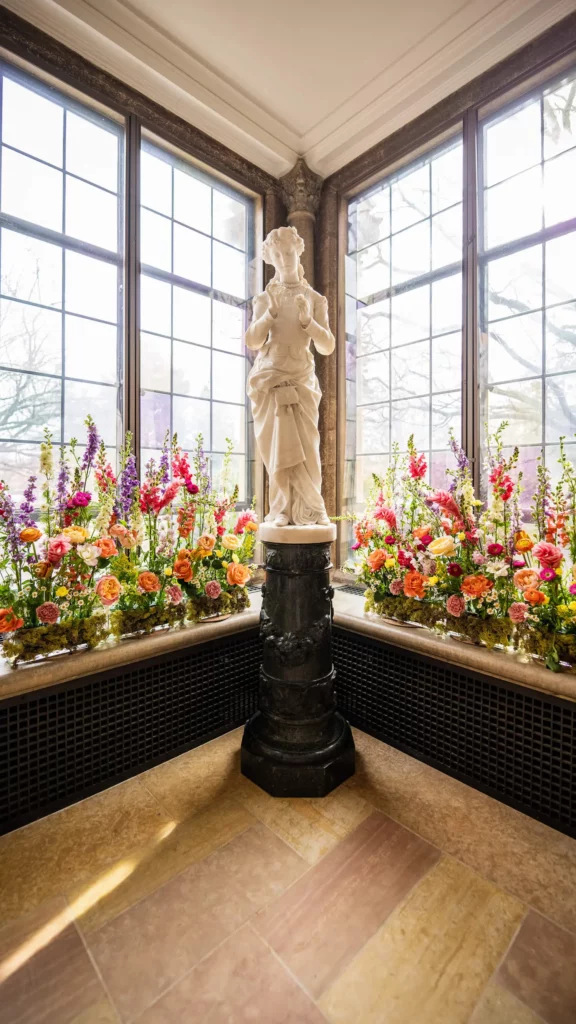 A statue “Margeurite” is surrounded by pink, orange, and green florals with stained-glass windows in the background.