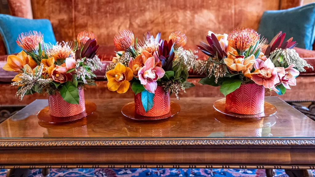 A coffee table in the Library is adorned with three floral arrangements featuring mauve, orange, green, and teal hues.