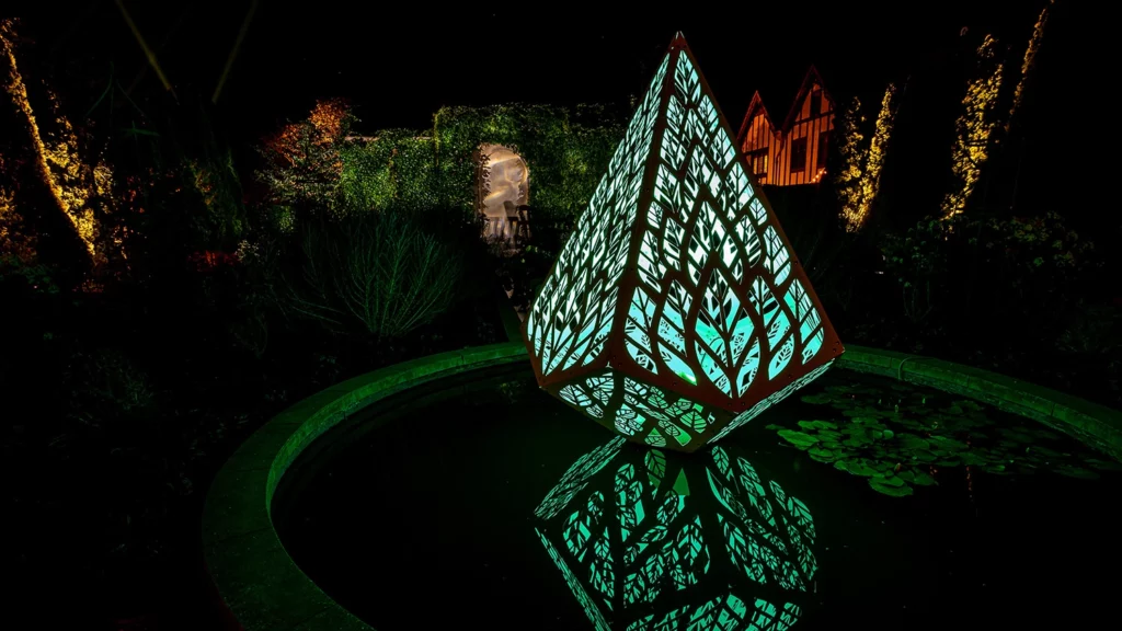 A pointed geometric sculpture illuminated in green light rests on top of a reflective pool and reflects shapes in the water.