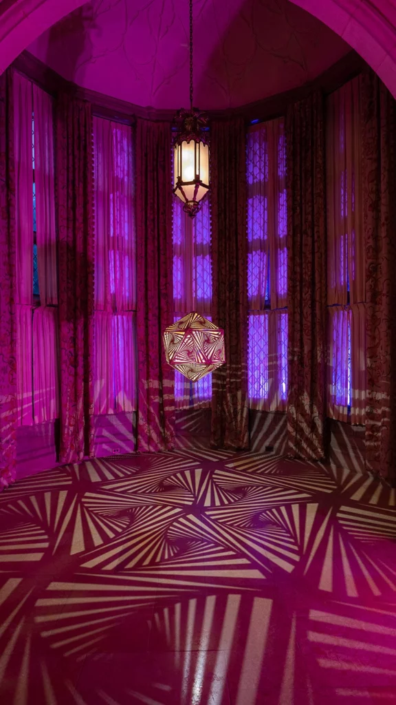 A rounded geometric sculpture illuminated with pink and yellow lighting hangs in the center of an alcove, casting shadows all around it.