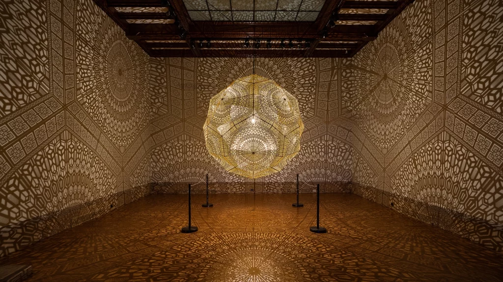 A large geometric sculpture hanging from the ceiling and bursting with yellow light reflects shapes all over the walls of a gallery.