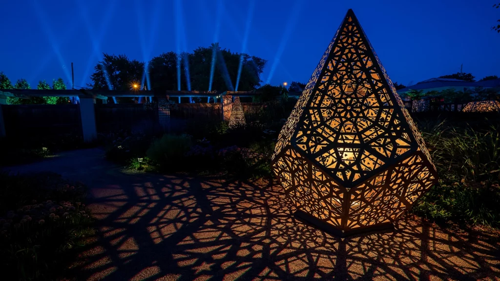 A pointed geometric sculpture illuminated in yellow lighting casts shape on the ground of a garden area.