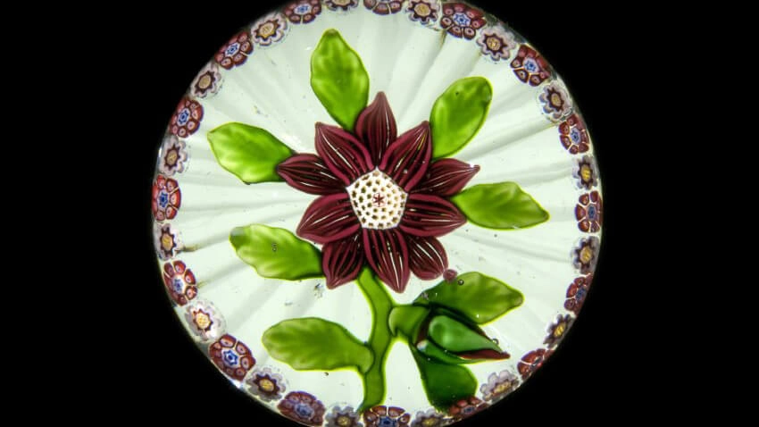 A paperweight with a maroon flower on a green vine, with maroon, white, and blue flowers along the circumference.