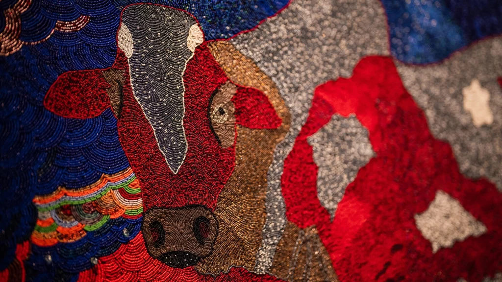 A close up of a beaded artwork, or “ndwango,” featuring a bull in shimmering red, silver, brown, and blue.