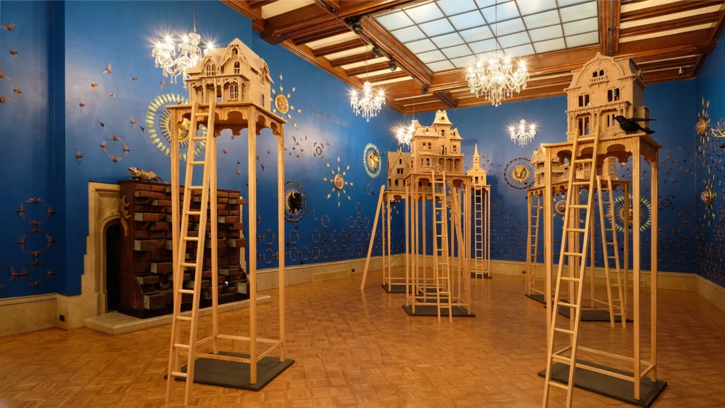 Wax house sculptures tower on stilts in the middle of the Main Gallery surrounded by insects pinned to bright blue walls in symmetrical patterns. A large, dresser of specimens sits at the forefront, and chandeliers dangle overhead.