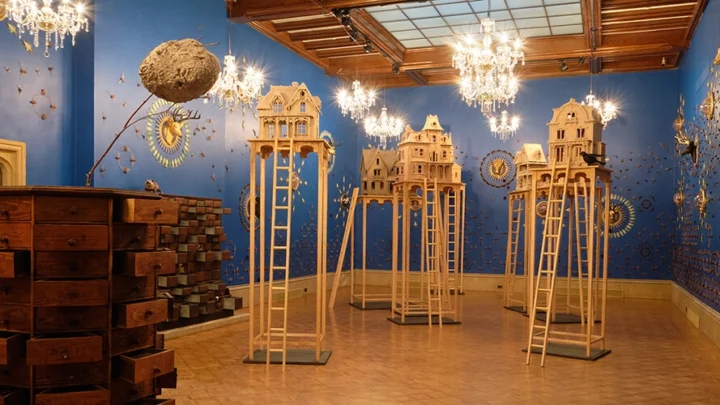 Wax house sculptures tower on stilts in the middle of the Main Gallery surrounded by insects pinned to bright blue walls in symmetrical patterns. A large, dresser of specimens sits at the forefront, and chandeliers dangle overhead.
