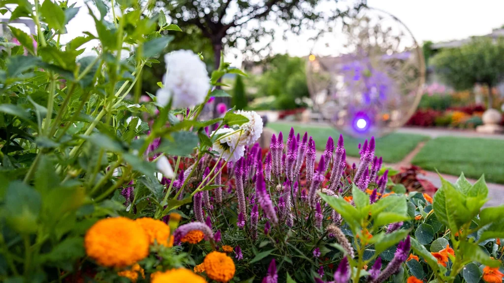 A metal geometric sculpture surrounded by blooming gardens.
