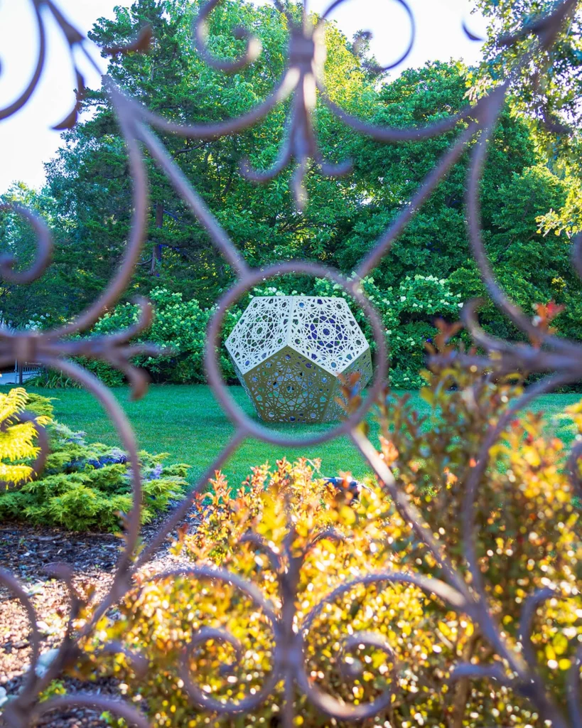 A metal geometric sculpture surrounded by blooming gardens.