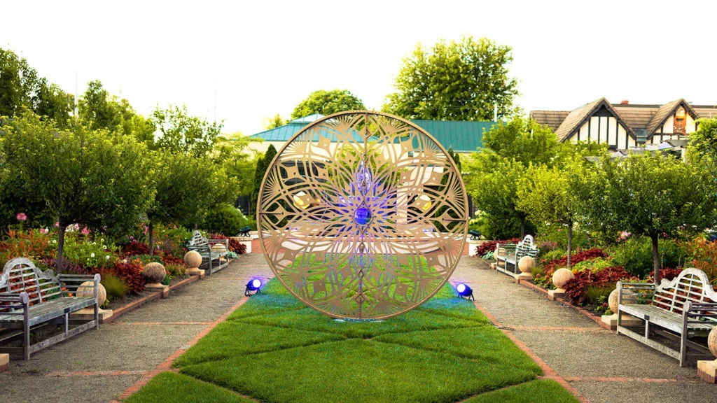 A metal geometric sculpture surrounded by blooming gardens.